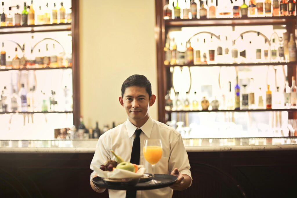 Waiter serving a drink and fruit tray in a bar with bottles displayed in the background.