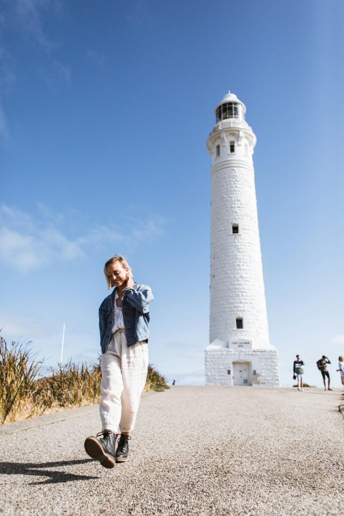 Full body of happy young female tourist adjusting blond hair while having stroll on road near majestic lighthouse in sunlight