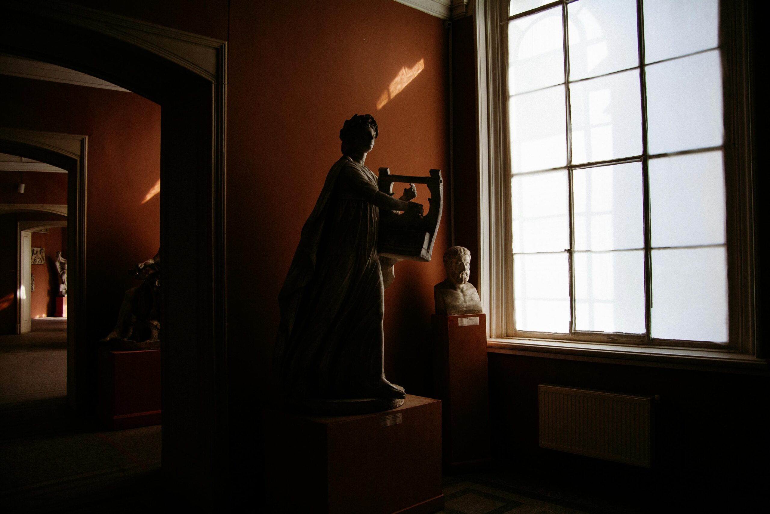 Classical sculptures illuminated by sunlight through a large window in a museum room.