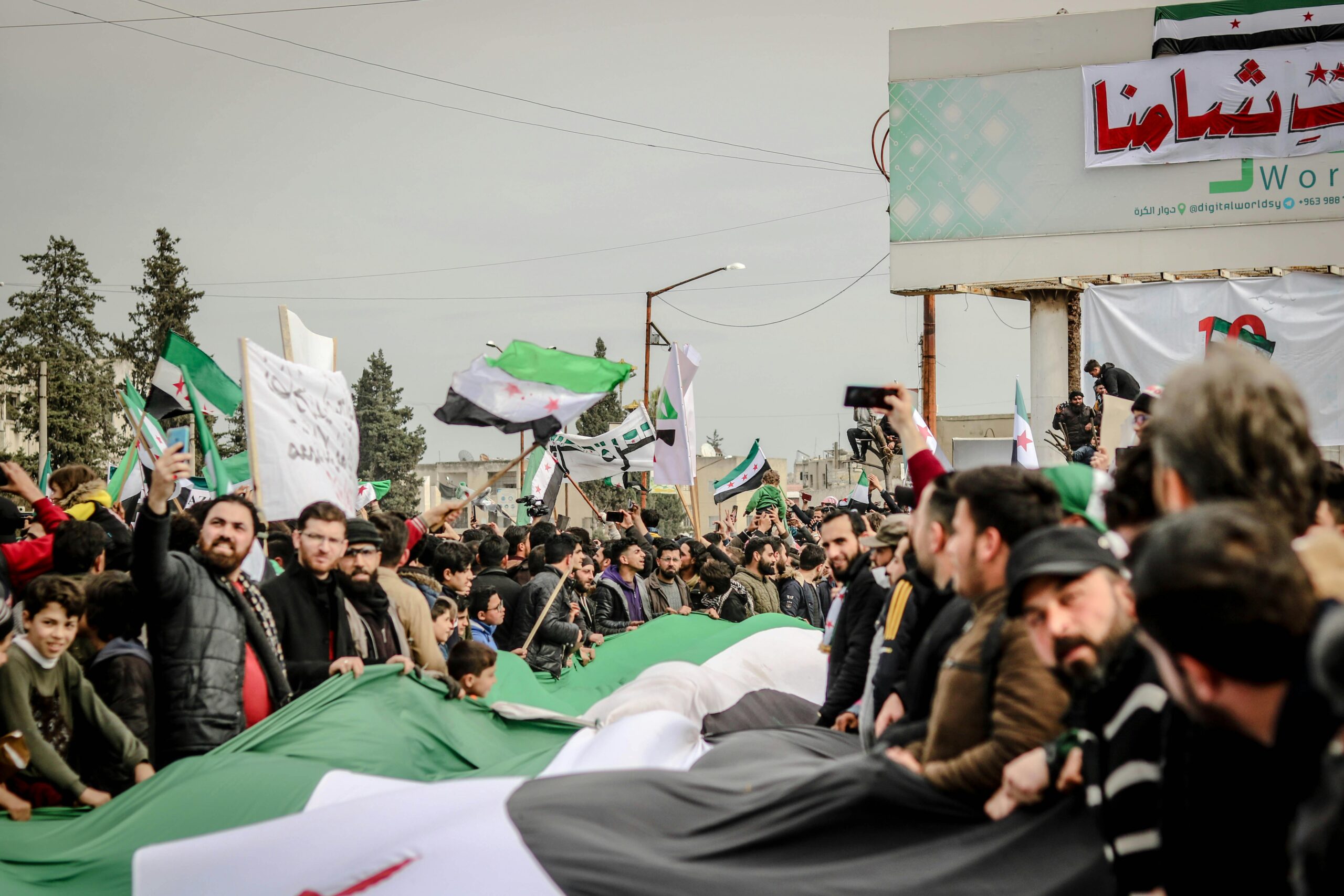 Crowds gather for a protest in Idlib, Syria, with flags and signs advocating political change.
