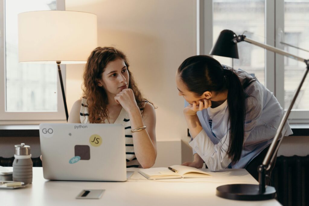 Two businesswomen collaborating on a project with a laptop and notebook in an office setting.