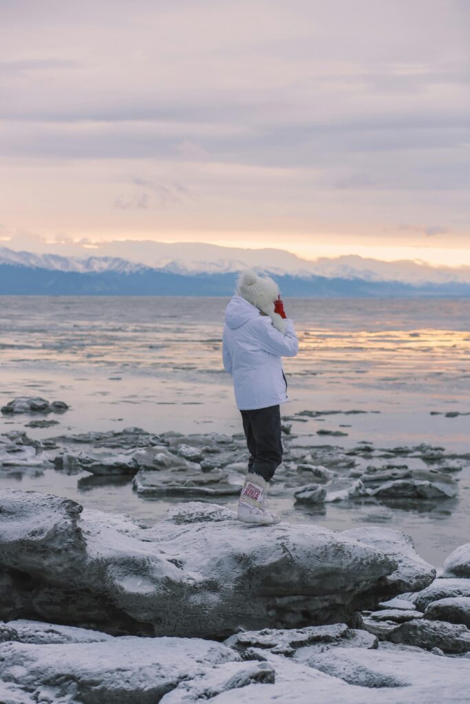 A traveler in winter clothing stands on ice by Anchorage, Alaska, enjoying a serene snowy landscape.