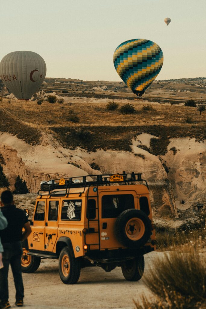 A rugged off-road vehicle parked in a rocky landscape with hot air balloons soaring above, creating a picturesque scene in Cappadocia.