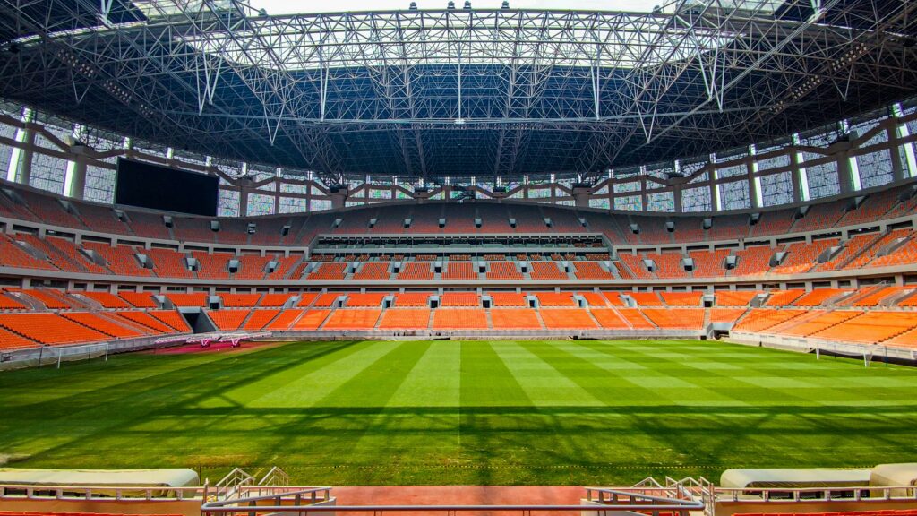 Panoramic view of an empty soccer stadium in Jakarta with vibrant orange seats and green field.