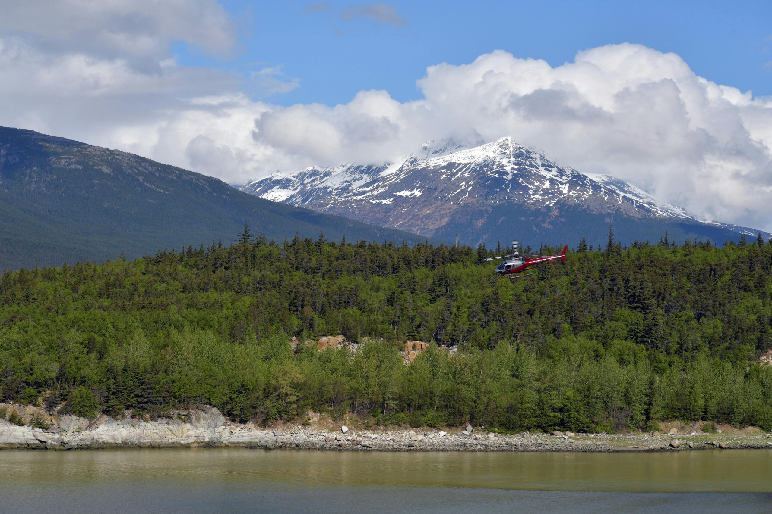 Helicopter flying over lush forests near Skagway, Alaska, with dramatic snow-capped mountains in the background.