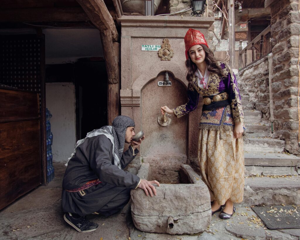 A man and woman in traditional Turkish outfits at a historic water well, showcasing cultural heritage.