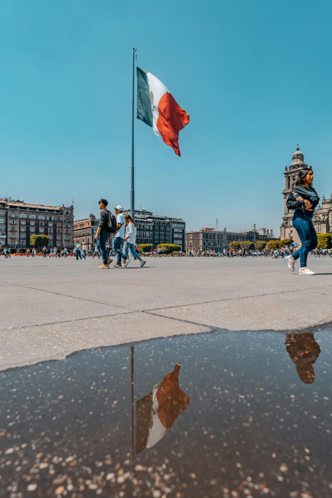 People walking through Plaza de la Constitución with the Mexican flag and cityscape in Mexico City.