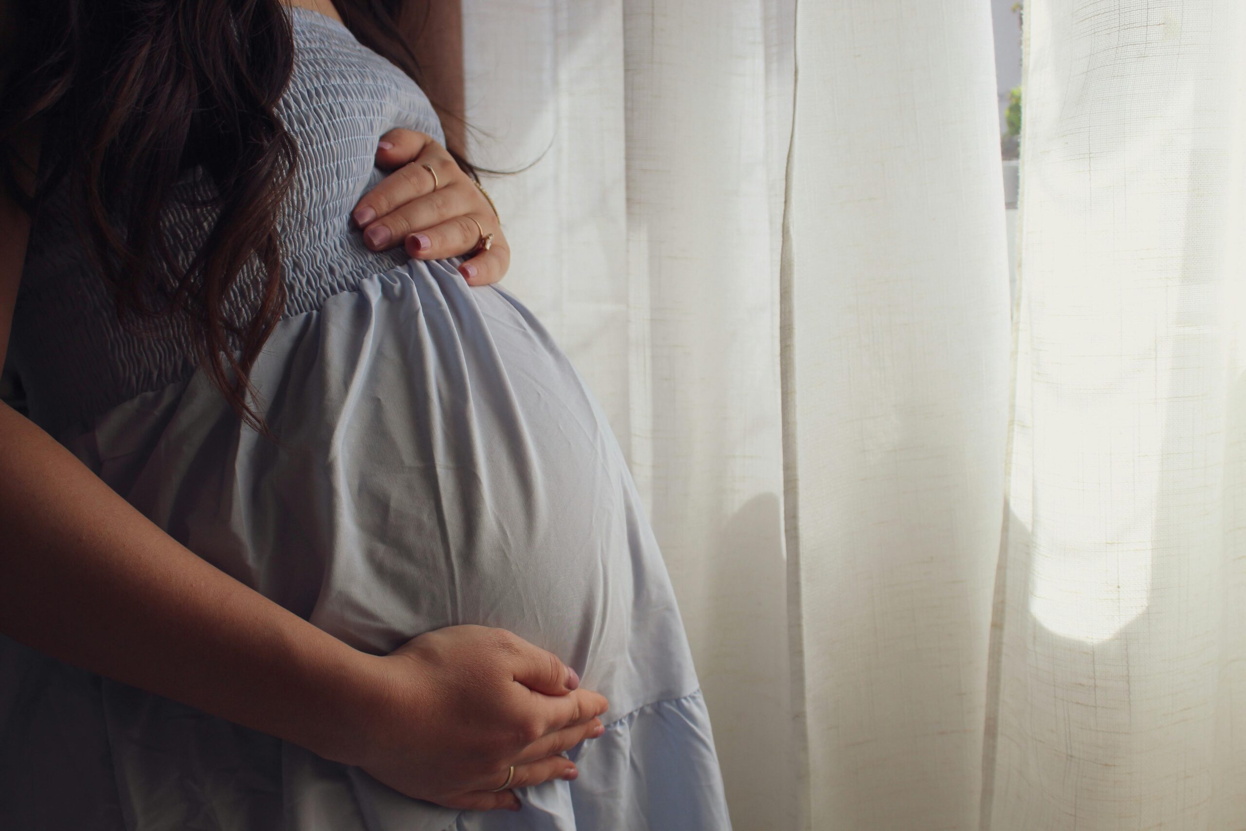A peaceful moment of a pregnant woman cradling her belly by a sunlit window indoors.