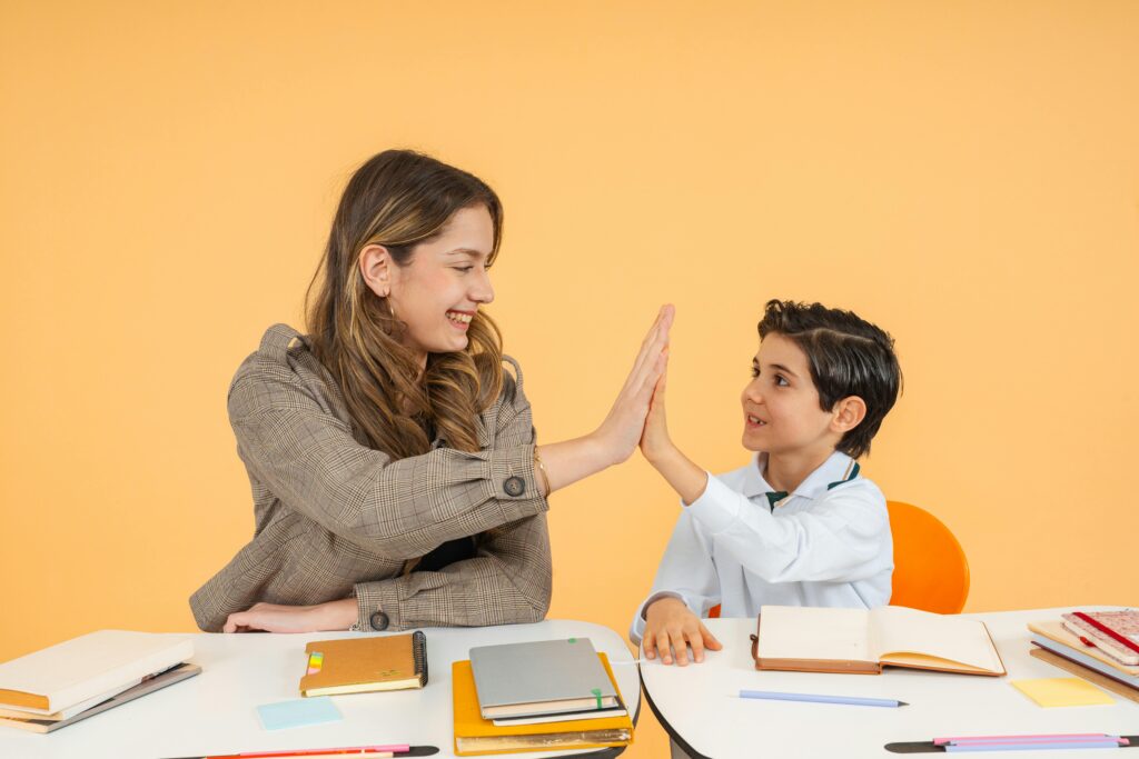 Teacher and student sharing a high-five over a desk in a bright classroom, fostering a positive learning environment.