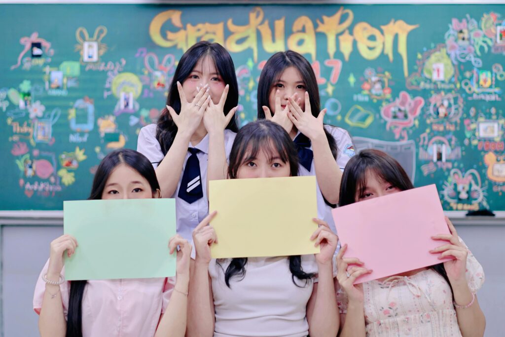 Five students hold colorful papers in front of a decorated blackboard during a graduation celebration.
