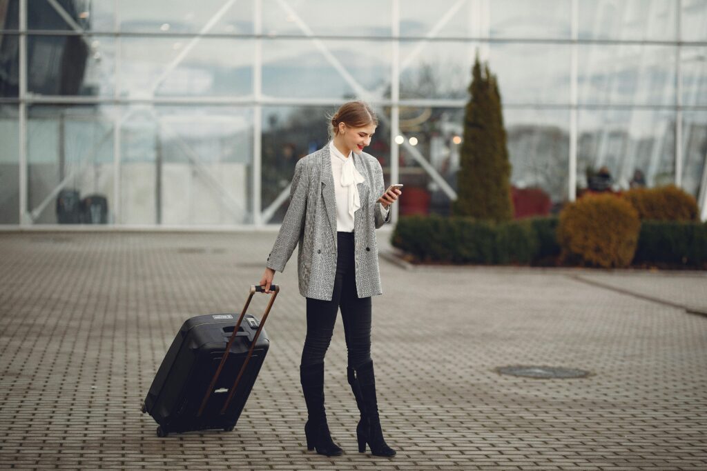 Full length of stylish woman in elegant outfit carrying baggage and surfing smartphone while standing near glass wall of airport