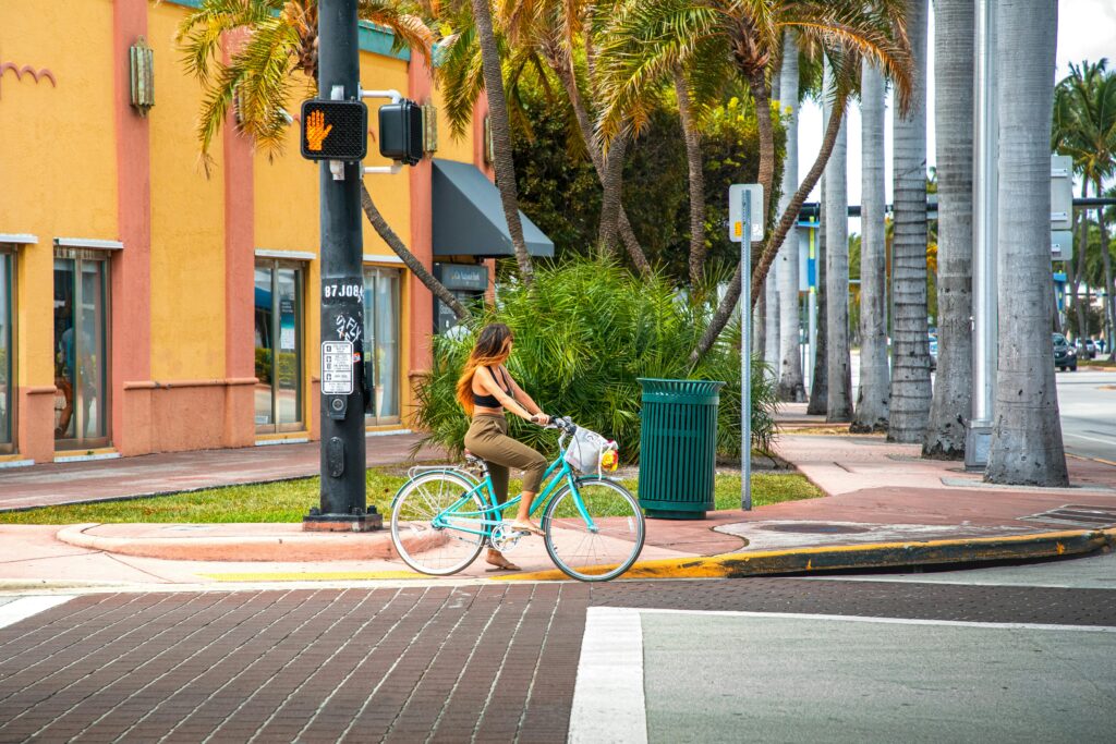 A woman rides a bicycle at a colorful Miami Beach street corner under palm trees.