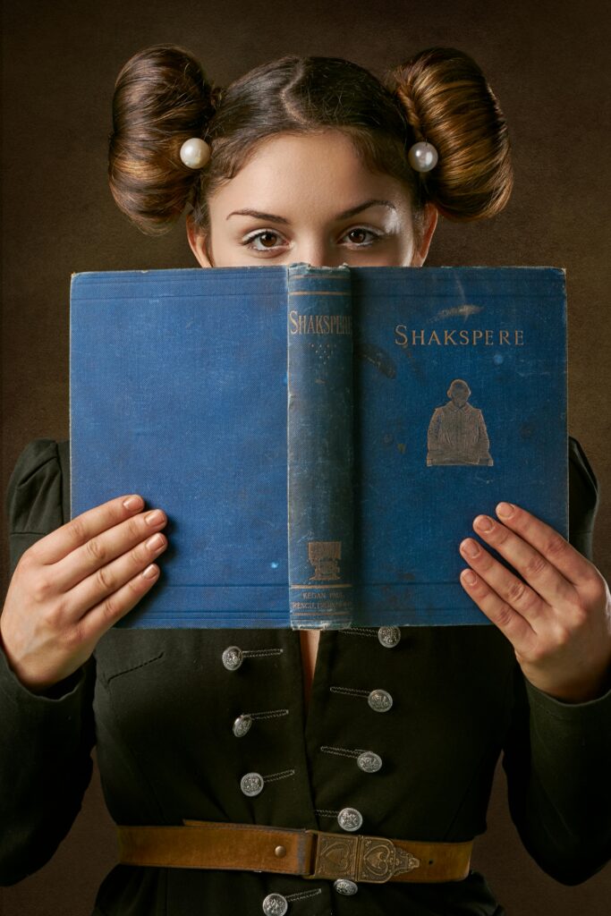 Young female dressed in medieval outfit with hair buns opened book of Old English poet in hands and looking at camera mysteriously