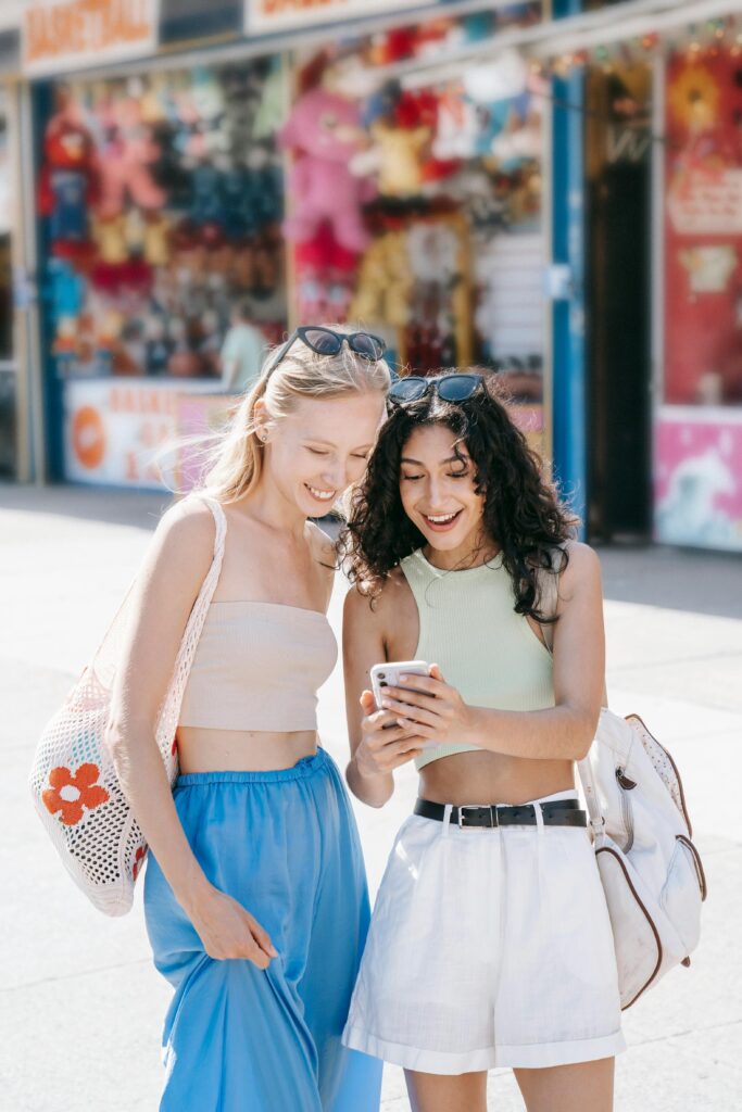 Two smiling young women enjoying a summer day, browsing on a smartphone outdoors.