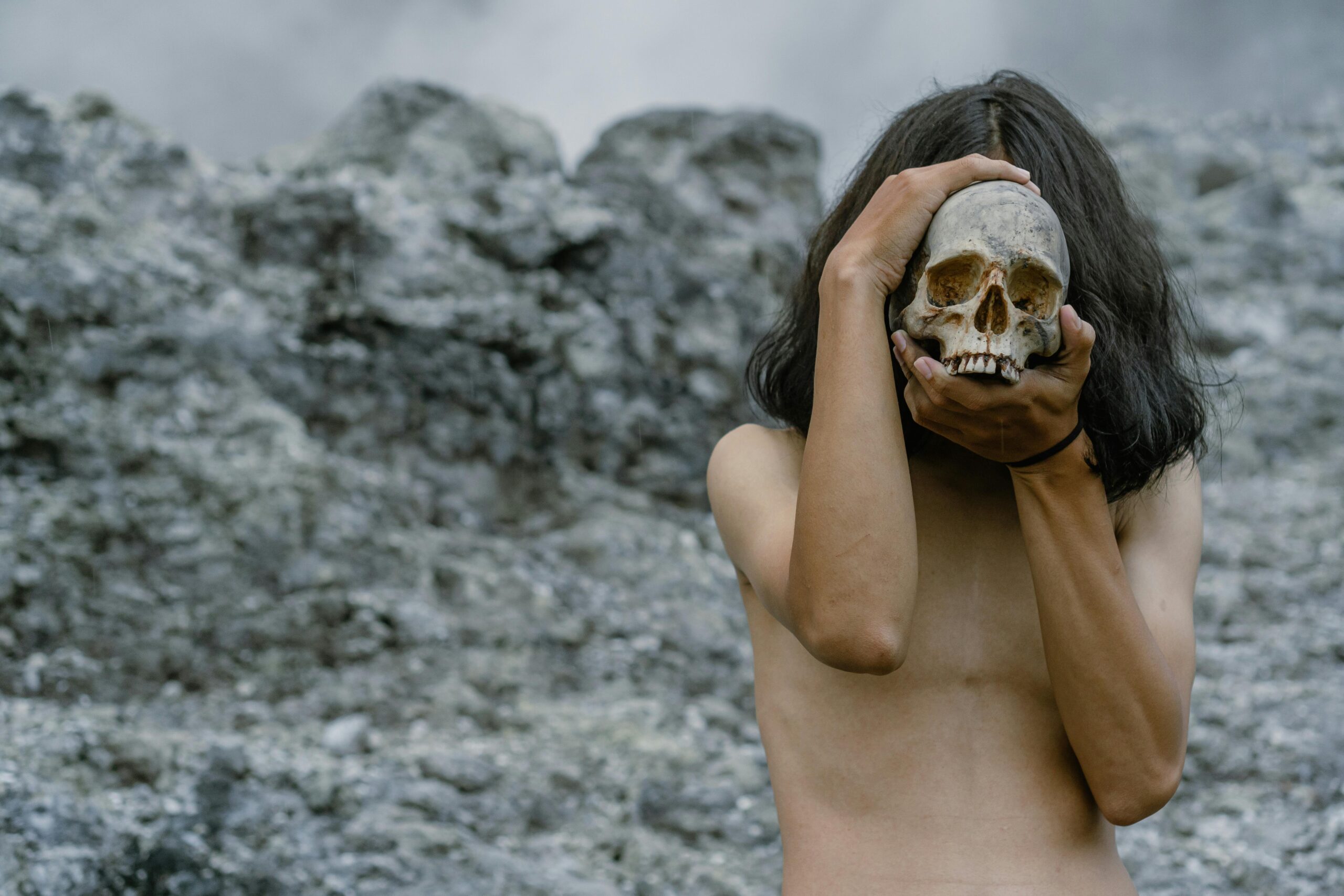 Topless individual holding a skull in a rocky outdoor environment, creating a mysterious and thought-provoking scene.