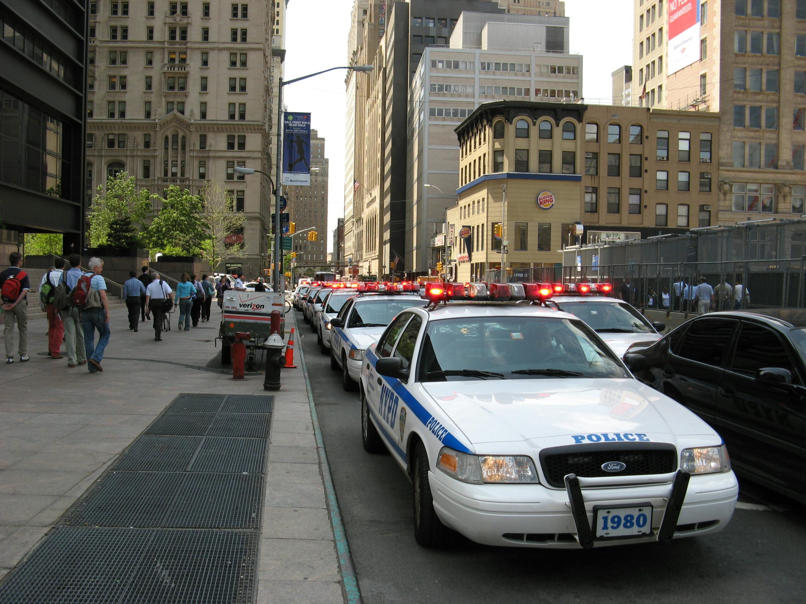 A line of NYPD police cars with flashing lights on a busy New York street, featuring pedestrians and tall buildings.