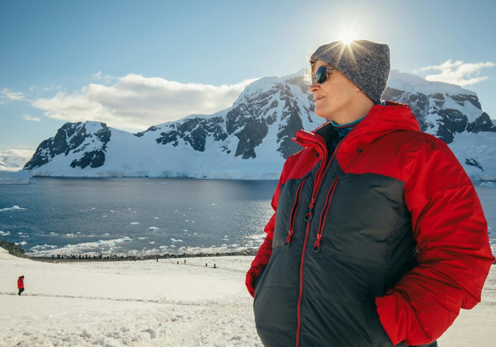 Man in red jacket enjoys scenic view of snowy Antarctic mountains and sea.
