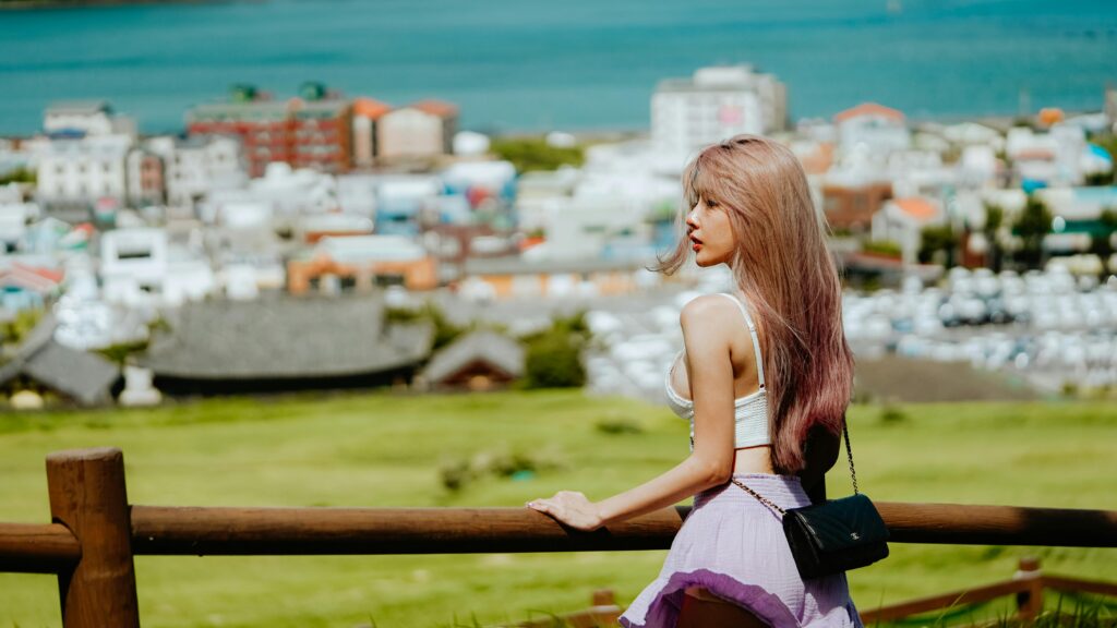 A fashionable woman with long hair views a scenic coastal town from a rustic fence.