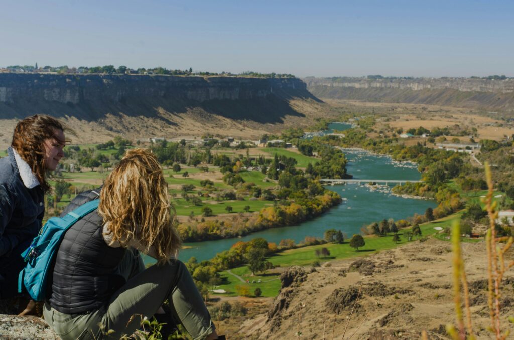 Two women sitting on a cliff enjoying a scenic view of a river valley with lush greenery.