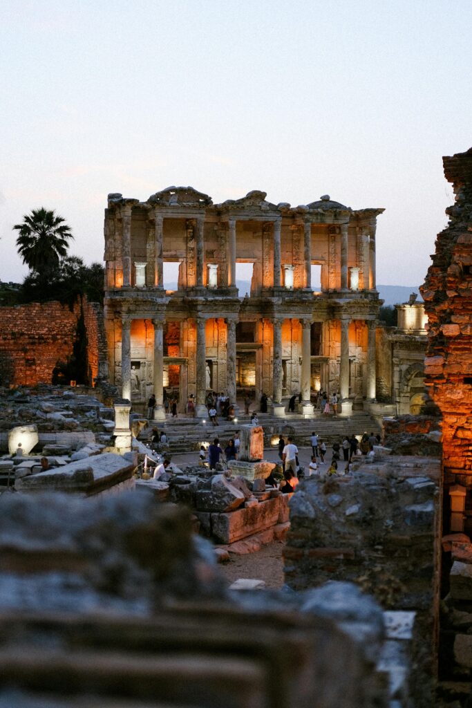 Illuminated Library of Celsus in Ephesus at sunset with tourists exploring the ancient ruins.