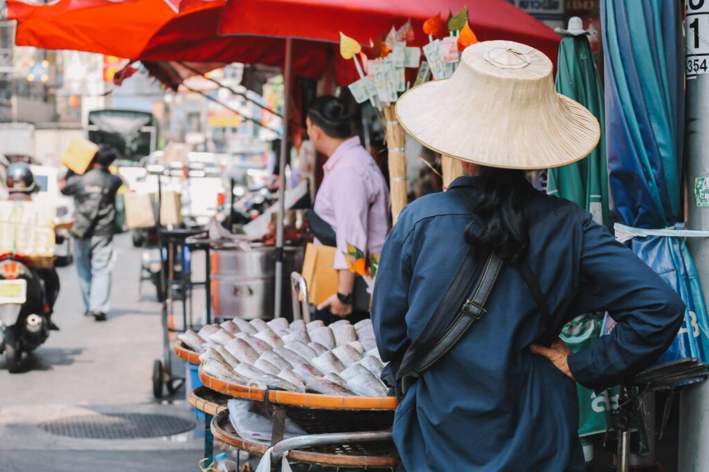 A busy Bangkok street market showing a vendor with fish under vibrant red umbrellas.