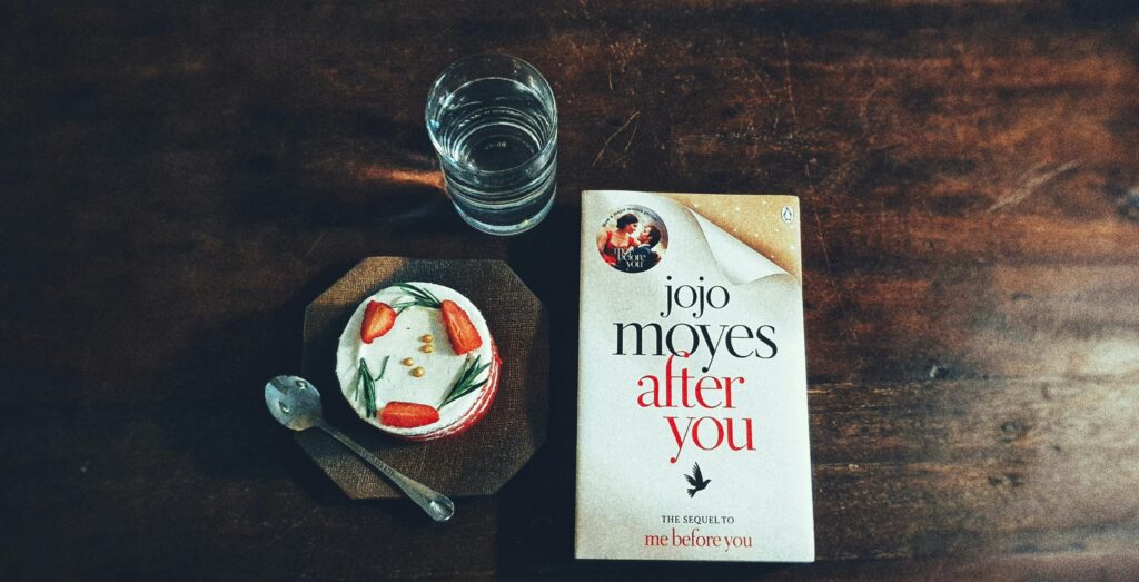 A cozy scene of Jojo Moyes' 'After You', a cake, and a water glass on a wooden table.