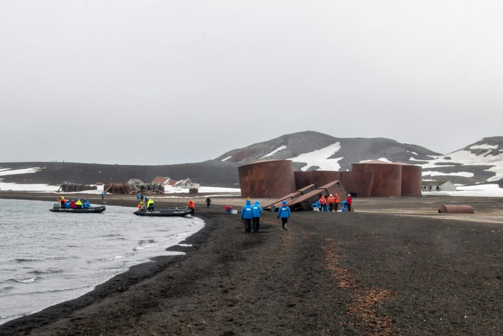 Explorers arrive by boat at Deception Island, Antarctica, with historical ruins and snowy landscapes.