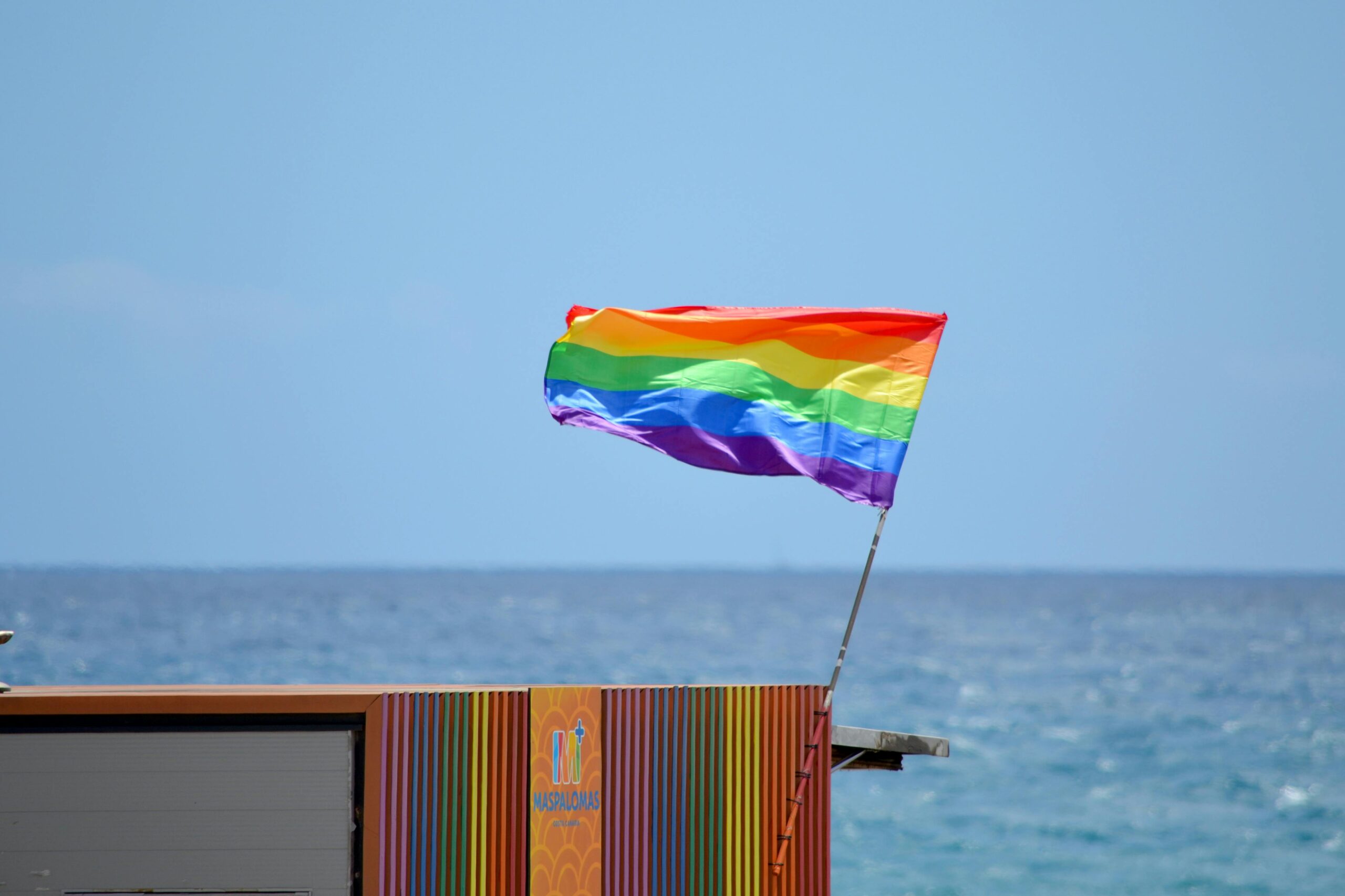 A vibrant rainbow flag waves at Maspalomas beach, symbolizing pride and diversity under the bright summer sky.