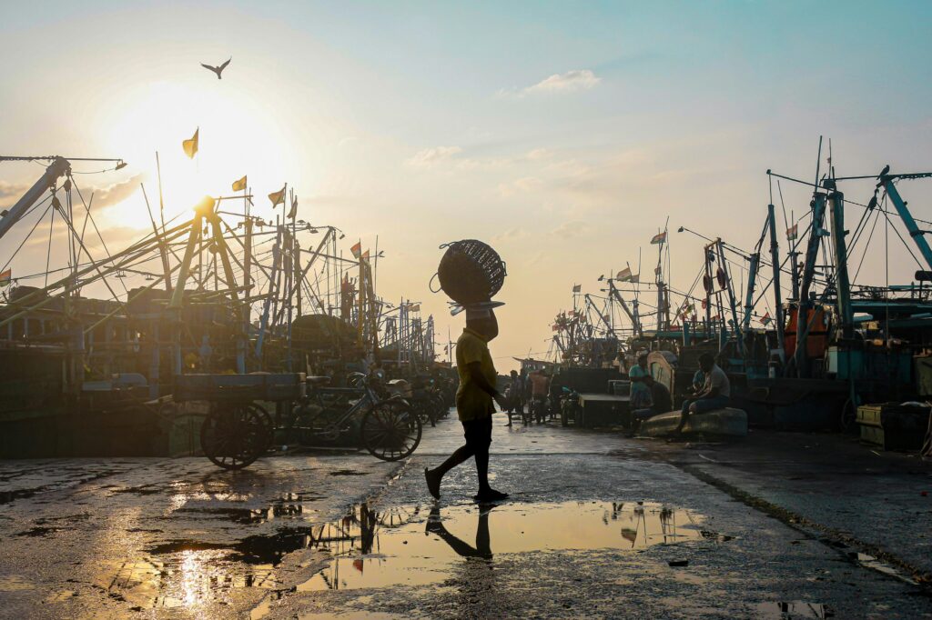 Silhouette of a fisherman carrying a basket in a bustling Chennai harbor at sunrise.