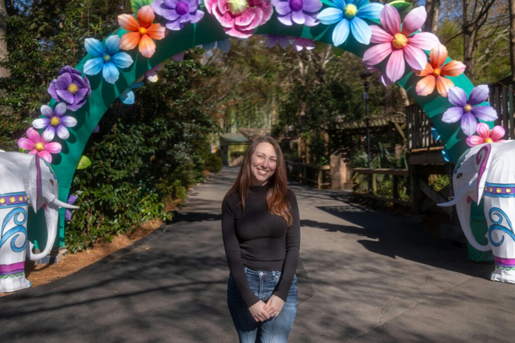 Happy woman standing under a colorful floral arch outdoors in a sunny park setting.