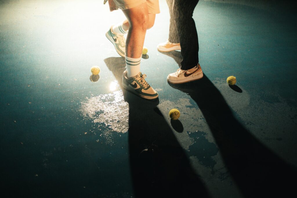 Close-up of tennis shoes and balls on wet court with dramatic lighting.