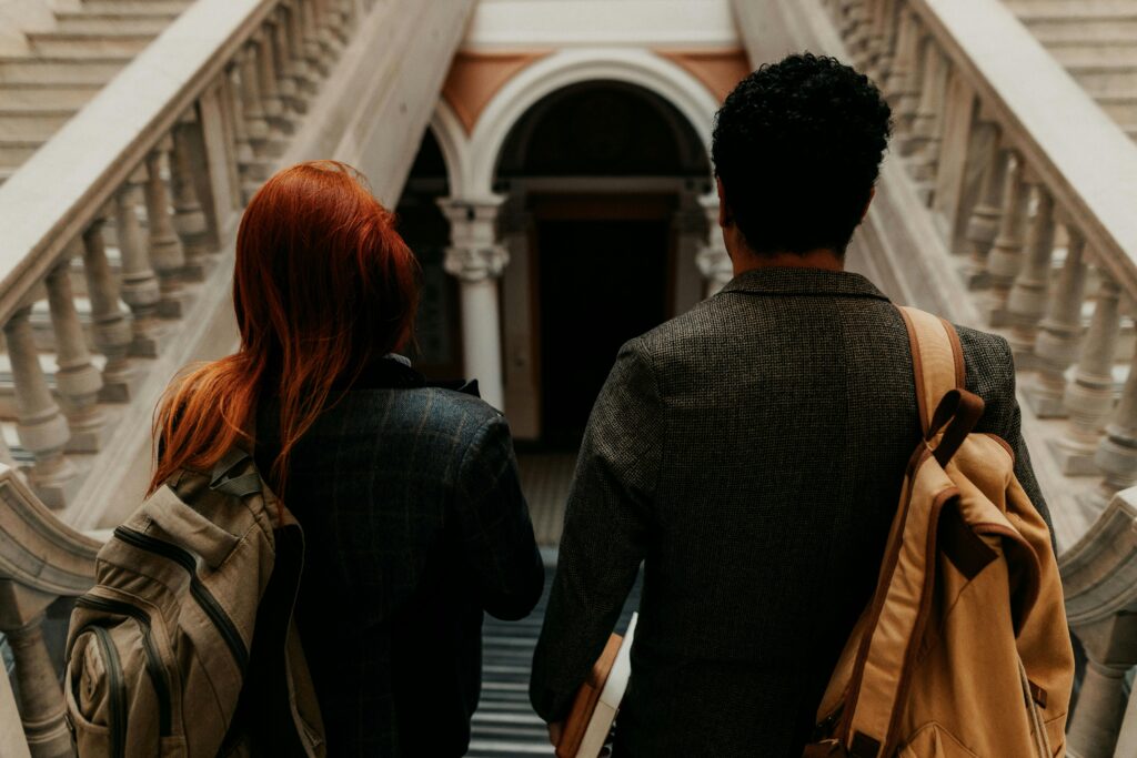 Two university students with backpacks walk down a grand staircase in a historic building.