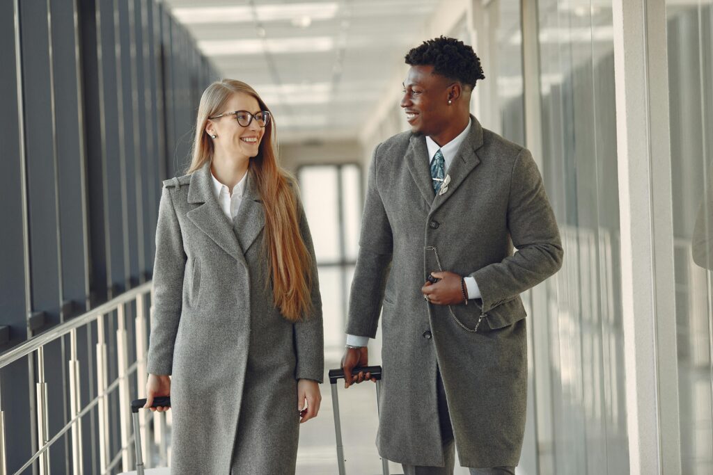 Positive smiling multiracial coworkers in classy formal clothing walking with luggage along modern glass airport airbridge in daytime and chatting