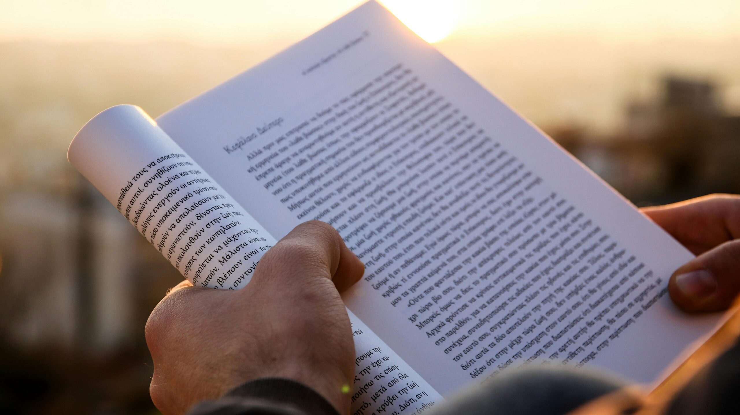 A close-up shot of hands holding a book with Greek text at sunset.