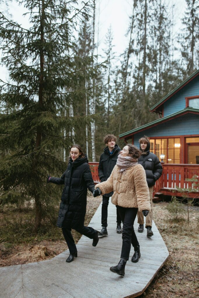 Young adults walk in winter coats along a forest path near a cozy cabin.