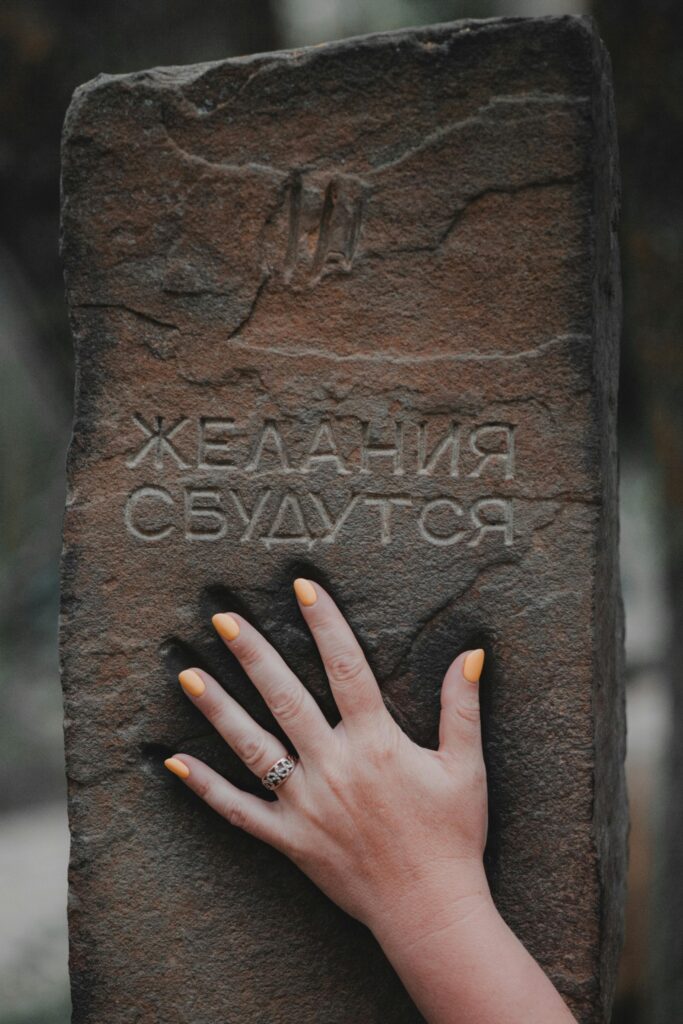 Stone slab engraved with Cyrillic text and a hand touching it, outdoor scene.