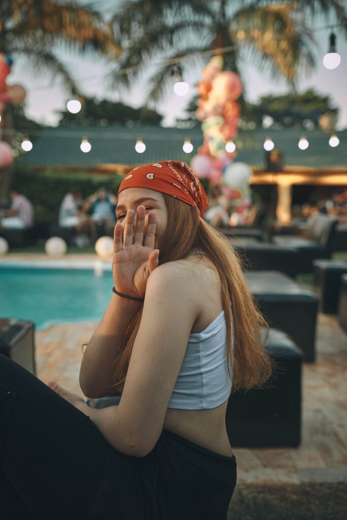 A young woman sitting by a pool, covering her face playfully with her hand, in a tropical outdoor setting.