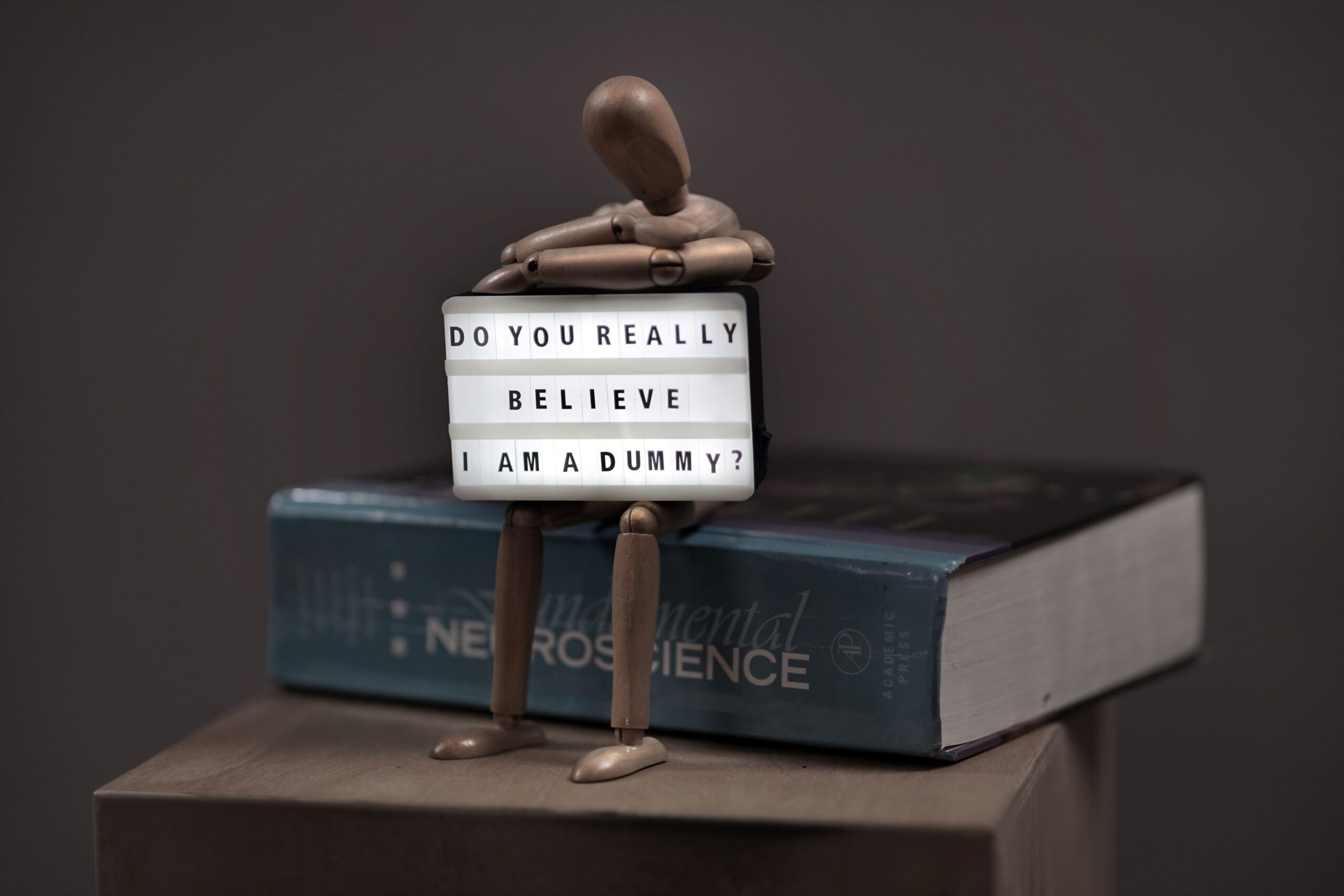 A wooden mannequin poses with a letter board reading 'Do you really believe I am a dummy?' atop a neuroscience book.