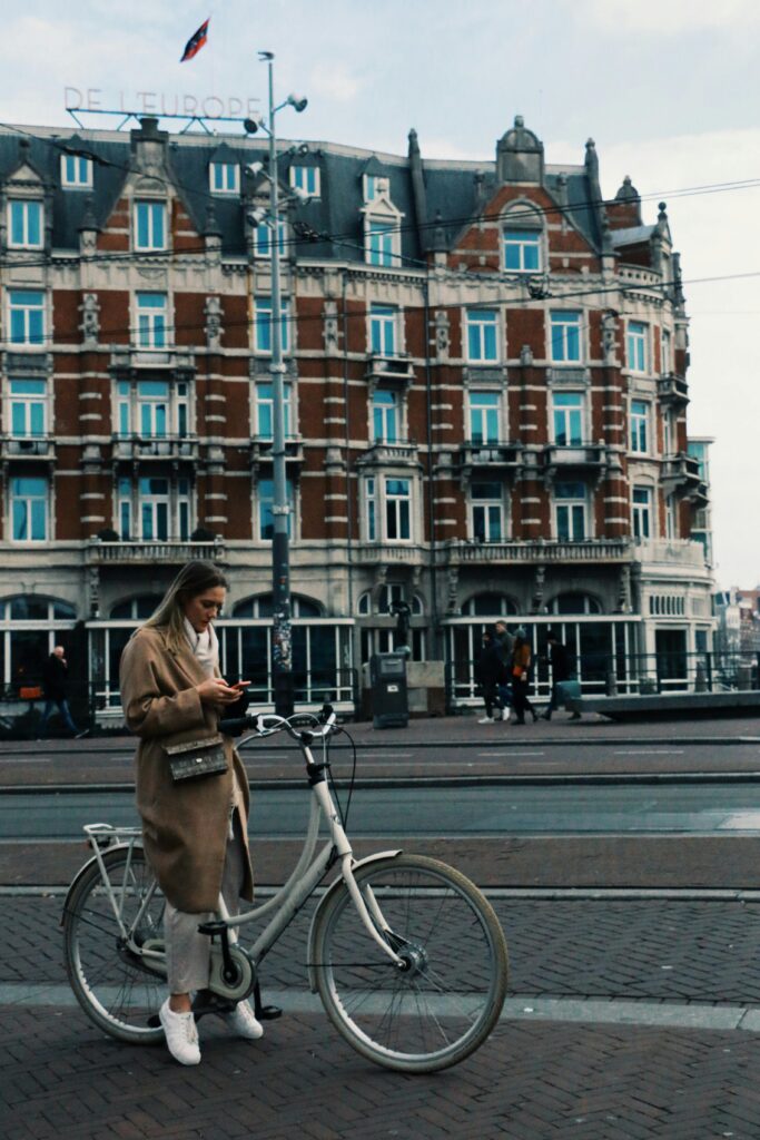 A woman stands with her bicycle on a European city street in front of a historic building.