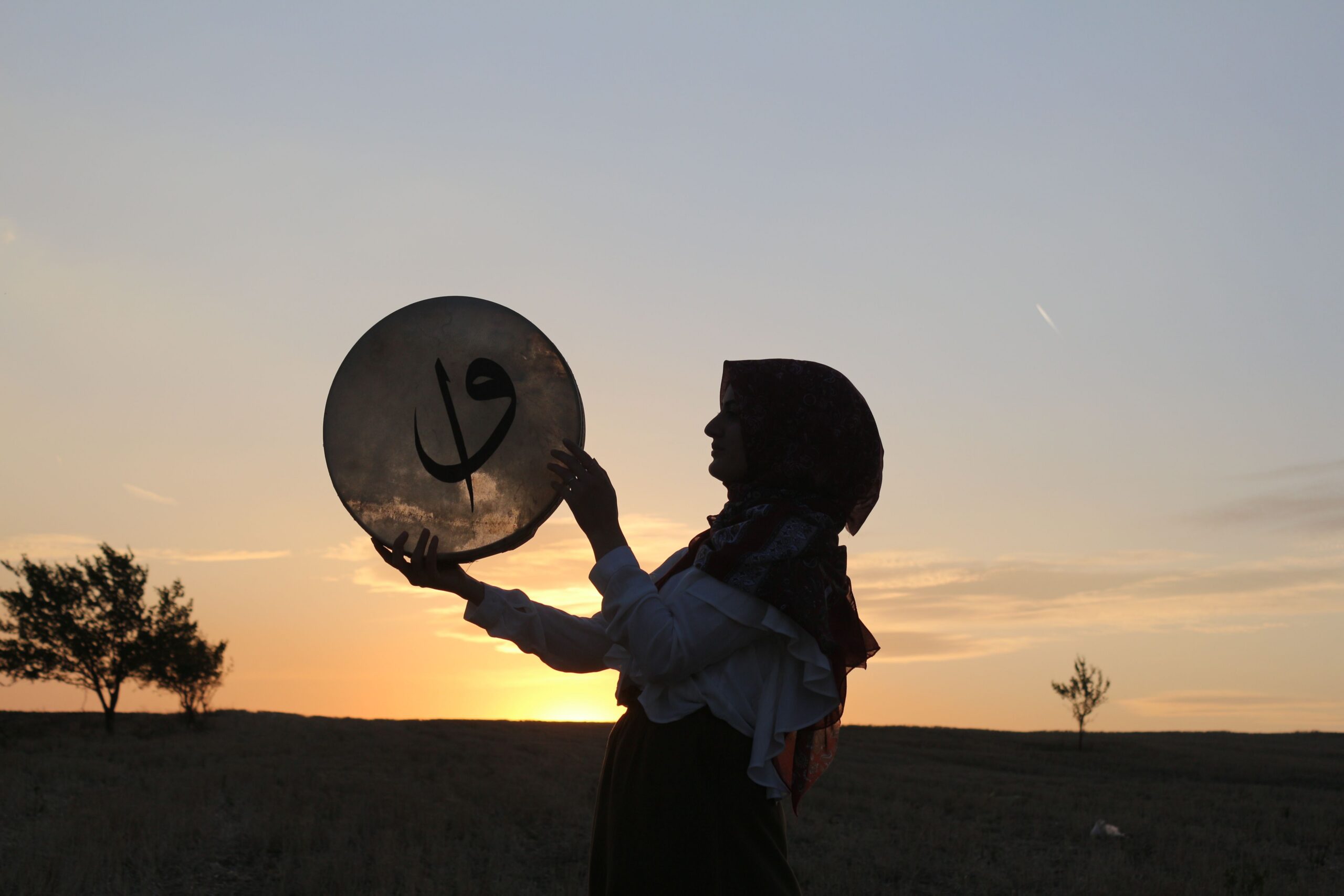 Silhouette of a woman holding a drum against a vibrant sunset in a rural landscape. Perfect for cultural or musical themes.