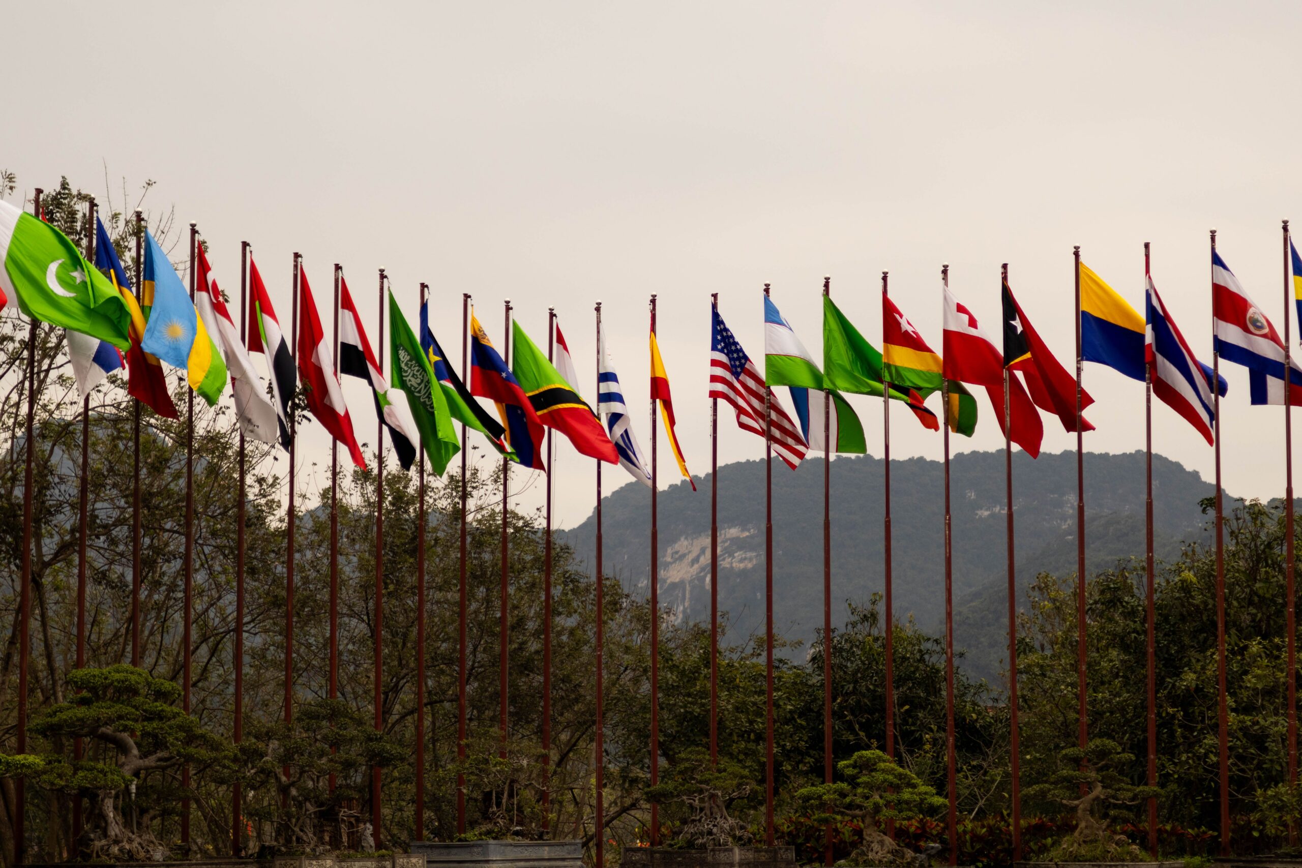 A display of various national flags on flagpoles set against a mountain backdrop, symbolizing global unity.