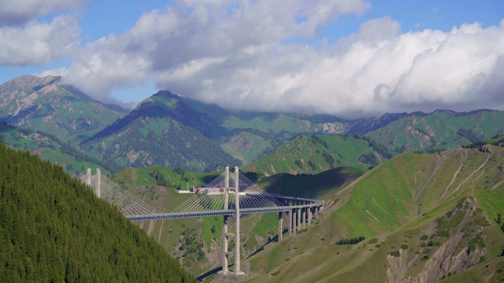 Scenic view of bridge spanning lush hills and Tianshan mountains under a cloudy sky.