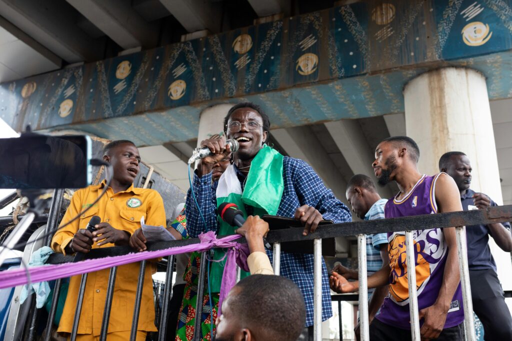 A lively public gathering with diverse participants singing and speaking under an urban bridge setting.