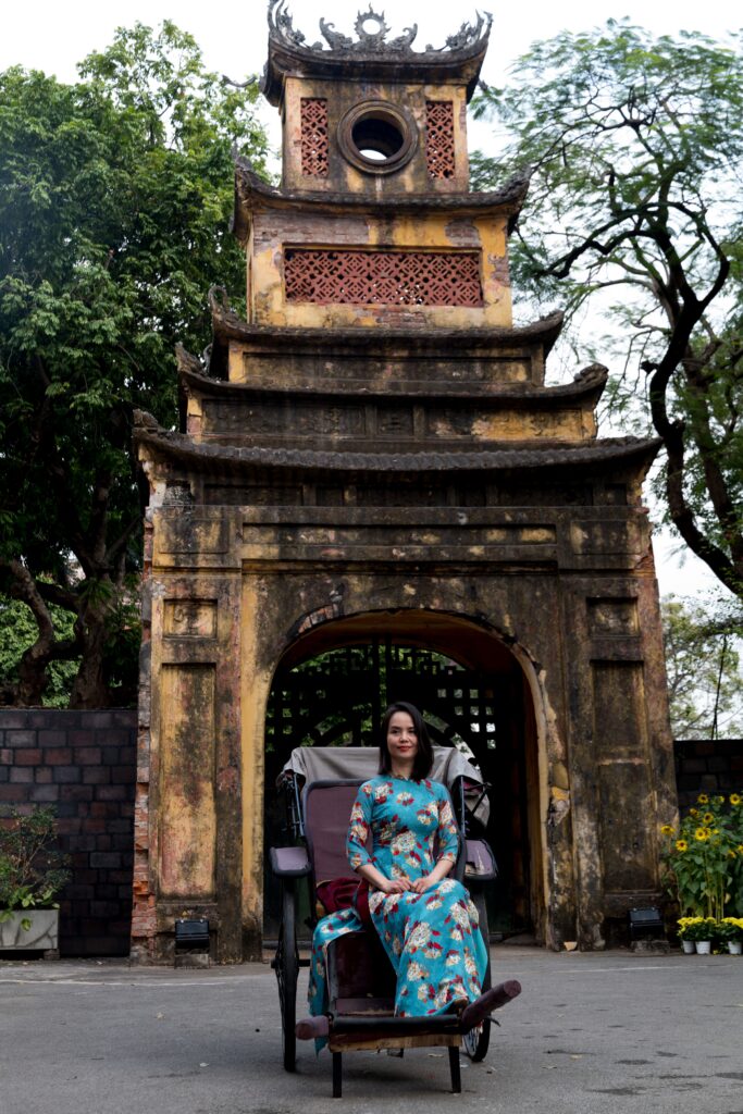 Woman in traditional attire sits in front of a historic gate in Vietnam, exuding cultural charm.