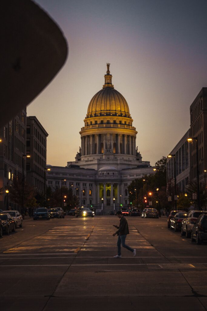 Person crossing street in front of the Wisconsin State Capitol at dusk. City lights illuminate the scene.
