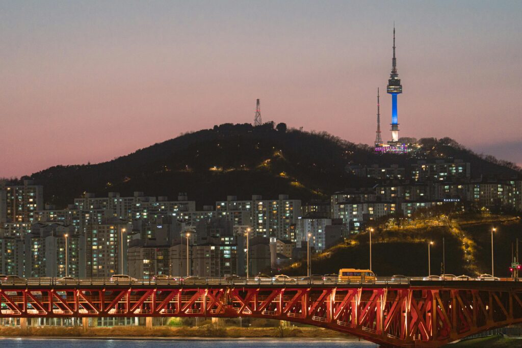 Seoul skyline at night with Namsan Tower illuminated, overlooking Han River and cityscape with a red bridge.