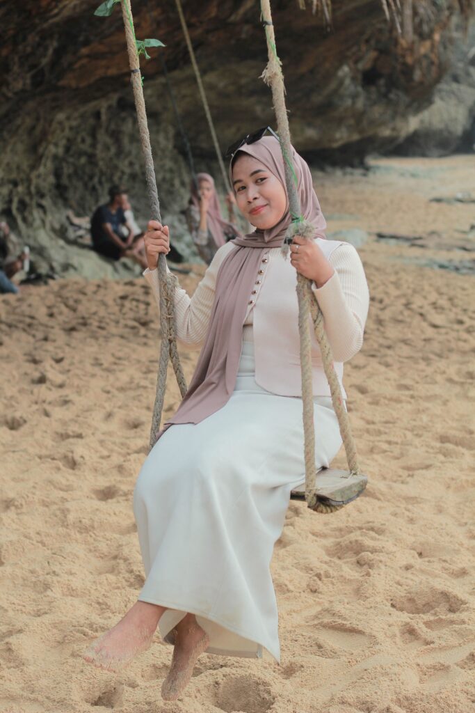 A woman in a hijab relaxes on a swing at a sandy beach with rocky cliffs.