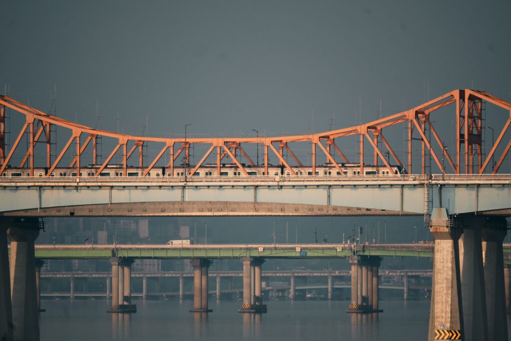 Train crossing an orange truss bridge above a river on a clear day.