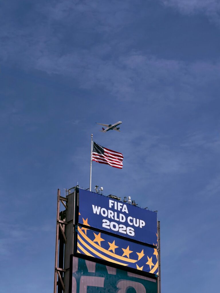 Airplane flying above FIFA World Cup 2026 sign with American flag, clear blue sky.