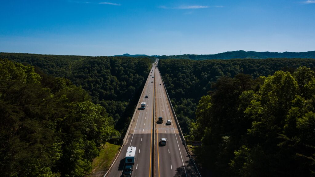 Aerial view of various vehicles riding on highway surrounded by lush green trees against blue sky on sunny day